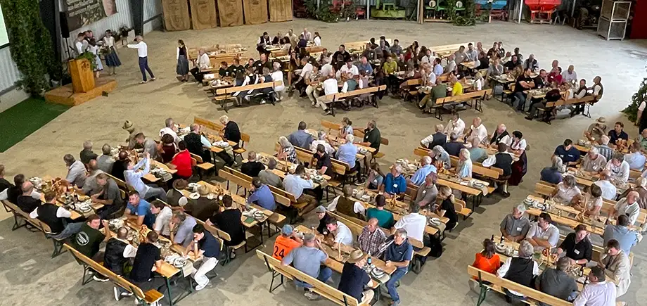 In einer landwirtschaftlichen Maschinenhalle sitzen ca. 200 Personen auf Bierbänken vor einer Bühne und einem Rednerpult.