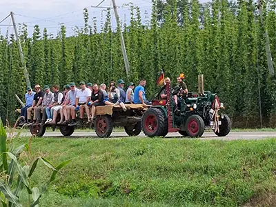 Vor einem grünen Hopfenfeld fährt ein Oldtimer-Tracktor. Auf dem offenen Anhänger sitzen viele Personen an den seitlichen Rändern und lassen die Beine nach unten baumeln.