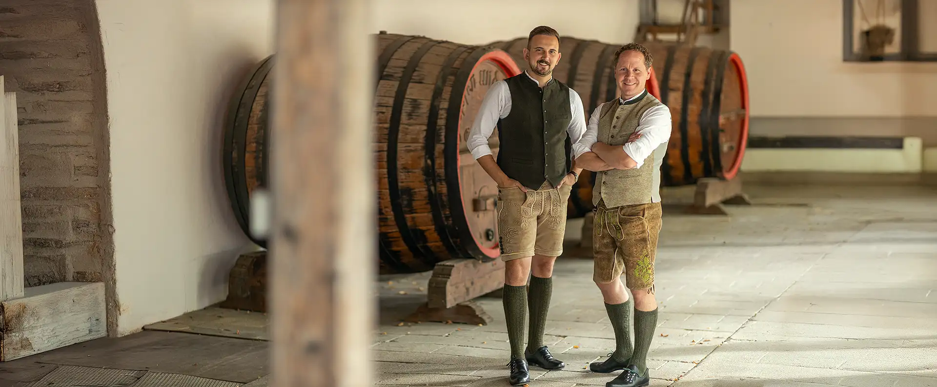 Christoph Goldbrunner und Mario Scholz, zwei Männer mittleren Alters in bayerischer Tracht gekleidet, stehen lächelnd vor 3 großen hölzernen Bierfässern